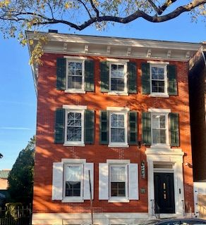 A brick building with green shutters, with a blue sky and tree branch in the background.